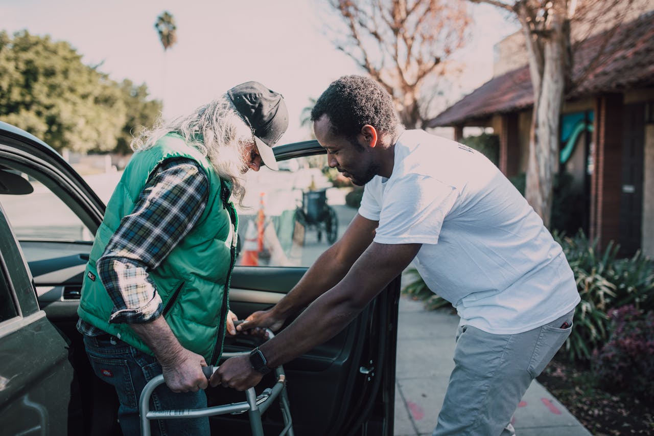 gallery-img-02 Young volunteer helping senior man with walker near a parked car, symbolizing care and support.
