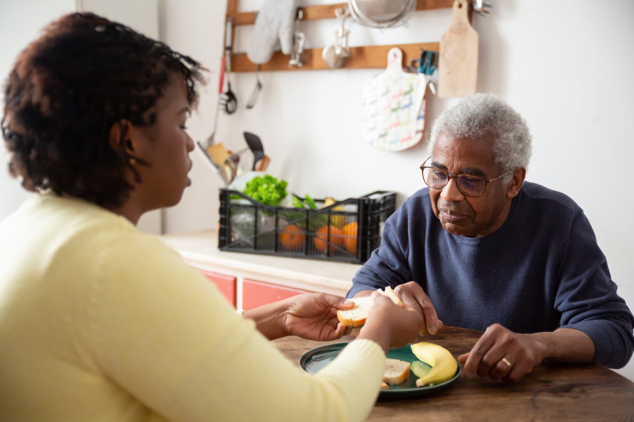 cta-01 A woman assists an elderly man with meals at home, showcasing care and support.