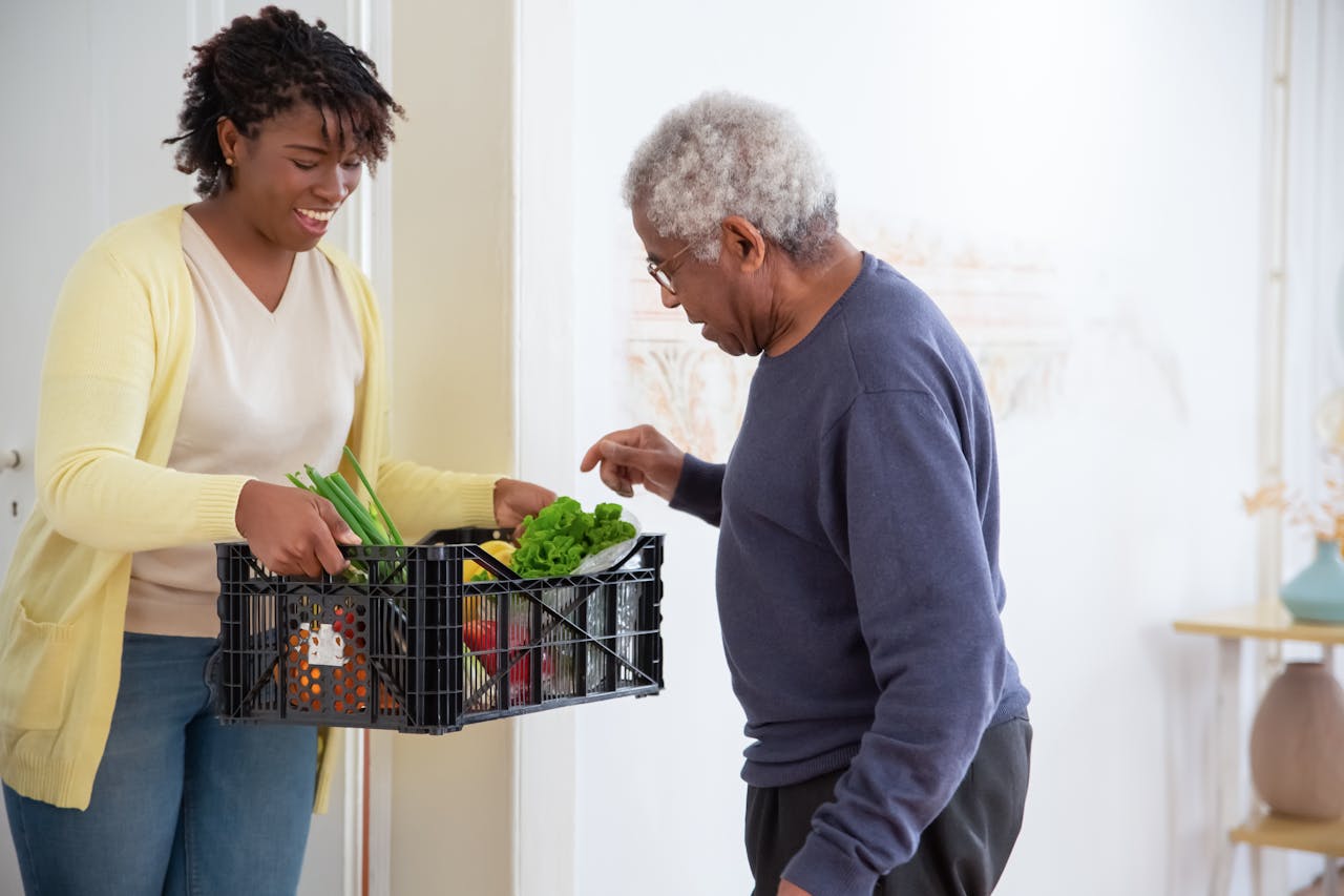 programs-03 A young adult woman helps a senior man by carrying a crate of fresh vegetables indoors, symbolizing care and support.