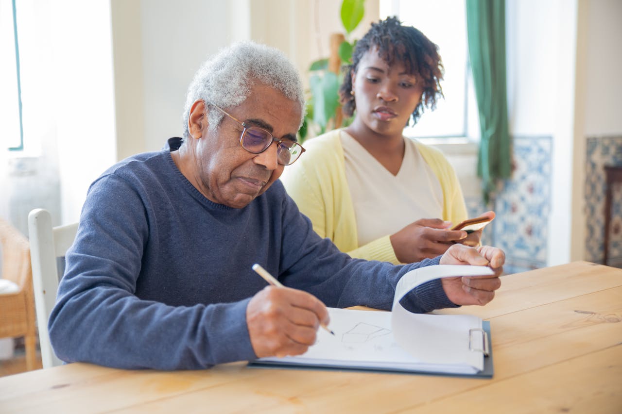 hero-gallery-03 A senior man writing on papers with a caregiver nearby indoors in a supportive setting.