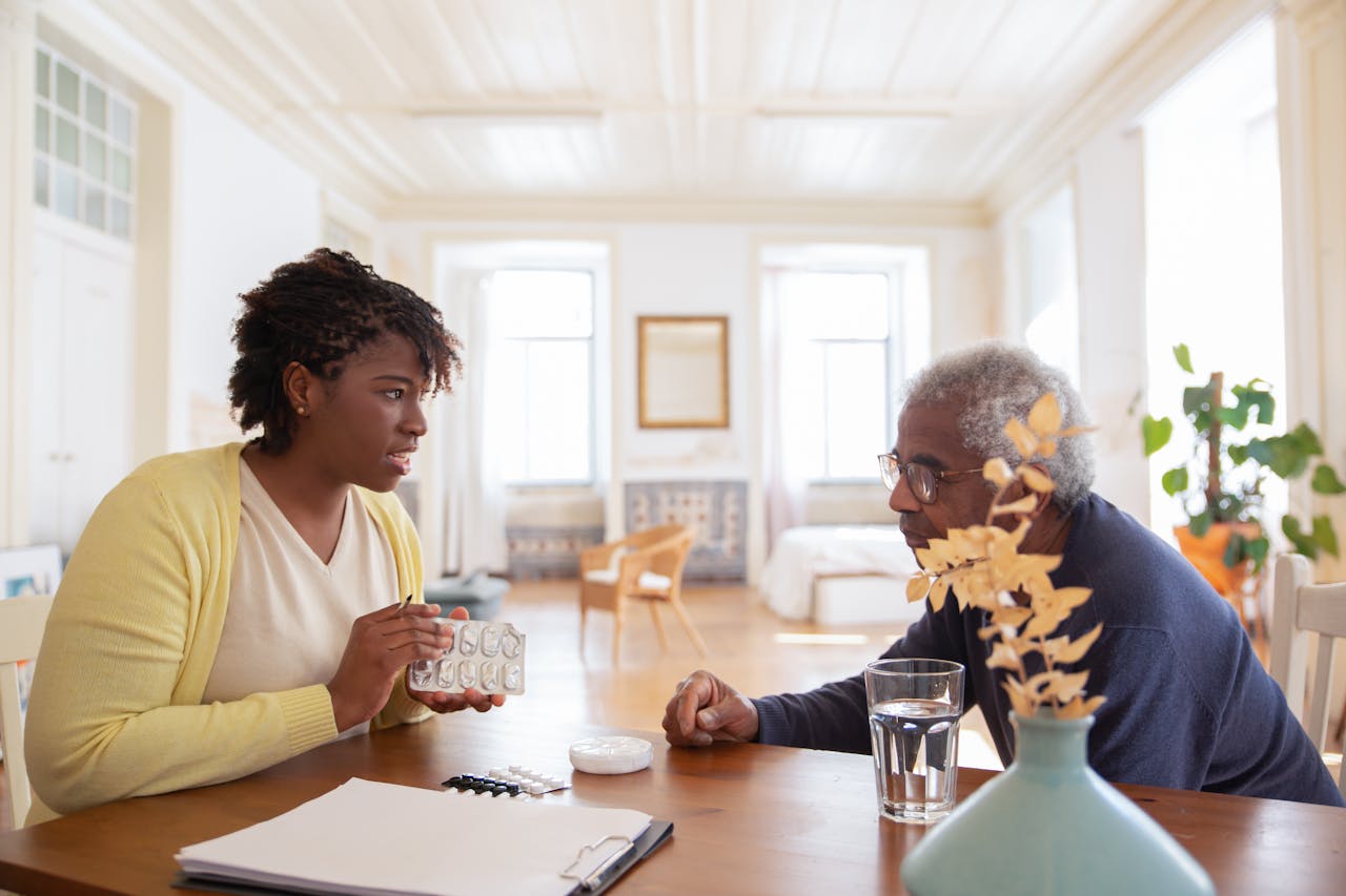 hero-gallery-04 An elderly man sits with a caregiver discussing medication at a table in a bright room.